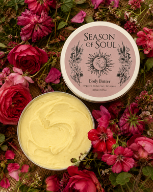 An open round tin of body butter showing pale yellow cream, with the matching lid placed beside it. The lid label reads 'Season of Soul' and 'Body Butter.