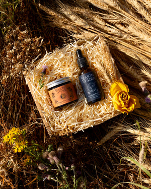 Two skincare products, a brown jar and a dark blue dropper bottle, arranged in a square woven basket filled with shredded paper on a bed of dried grass and flowers.