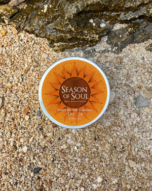 Tin of 'Season of Soul' sunscreen on a sandy surface with rocks.
