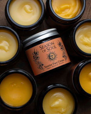 A jar of Season of Soul Sunrise Face Cream sits in the center, surrounded by multiple open black jars filled with a thick yellow balm, arranged on a dark textured surface.
