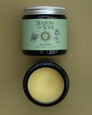 A jar of Season of Soul Repair Balm shown closed with a sage green label and silver lid, above an open jar displaying the pale yellow balm inside, on a solid olive background.