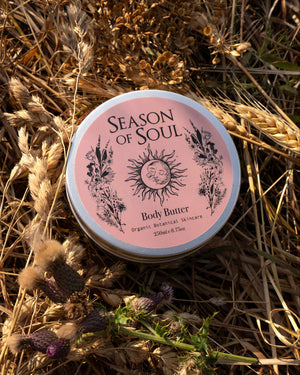 A round tin of Season of Soul Body Butter lies on a bed of dried wheat stalks and purple thistles. The lid is light pink with black text and illustrations of a sun and botanicals.