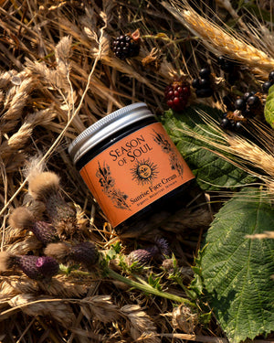 A jar of Season of Soul Sunrise Face Cream placed on a bed of dried wheat, thistles, and green leaves outdoors.