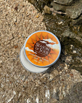 Tin of 'Season of Soul' sunscreen on a sandy surface with water and rocks.