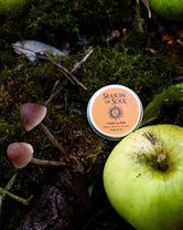 Tin of 'Season of Soul' product on a mossy surface with green apples and mushrooms.