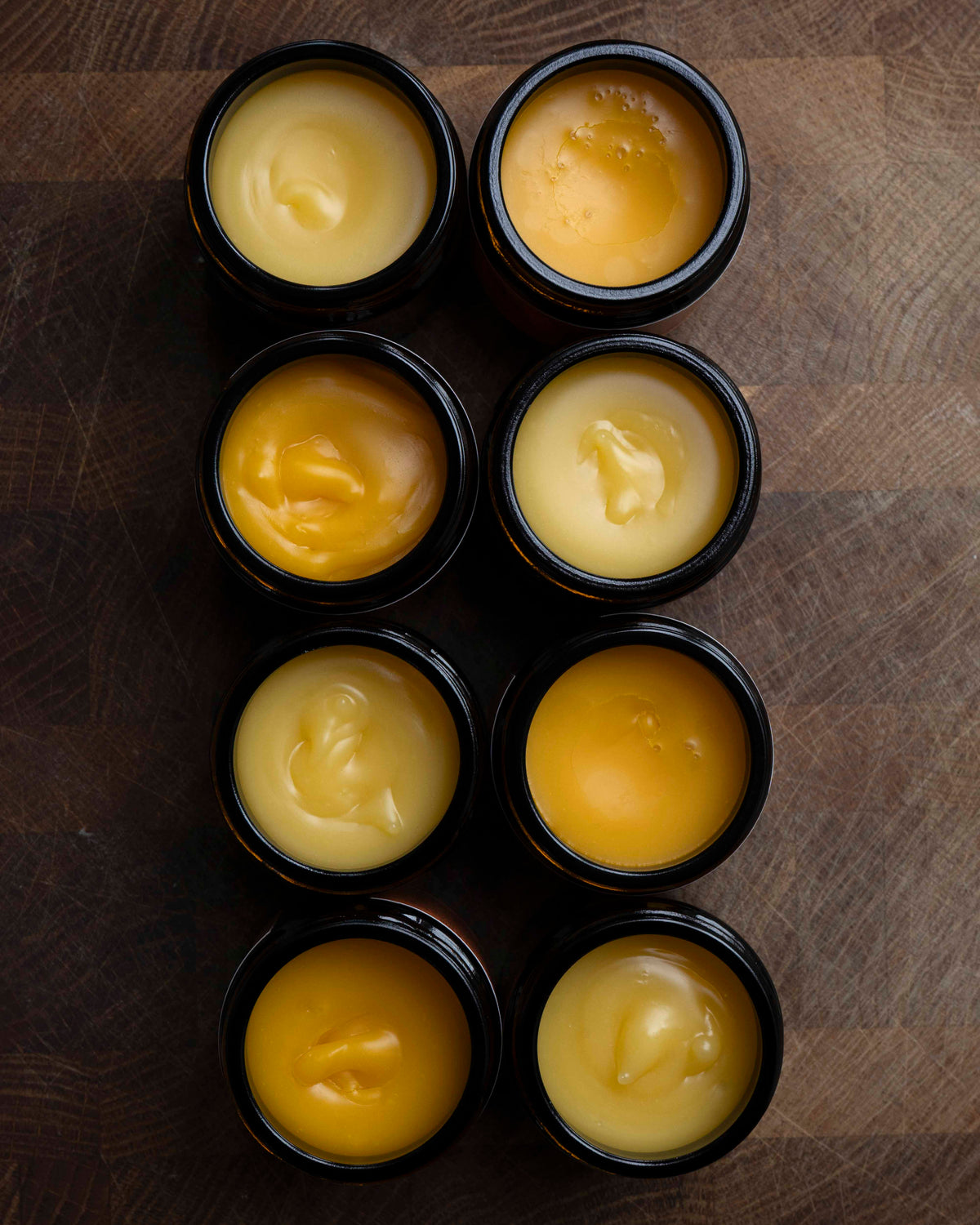 Eight small black jars filled with hand made face cream, arranged in two columns on a dark wooden surface.