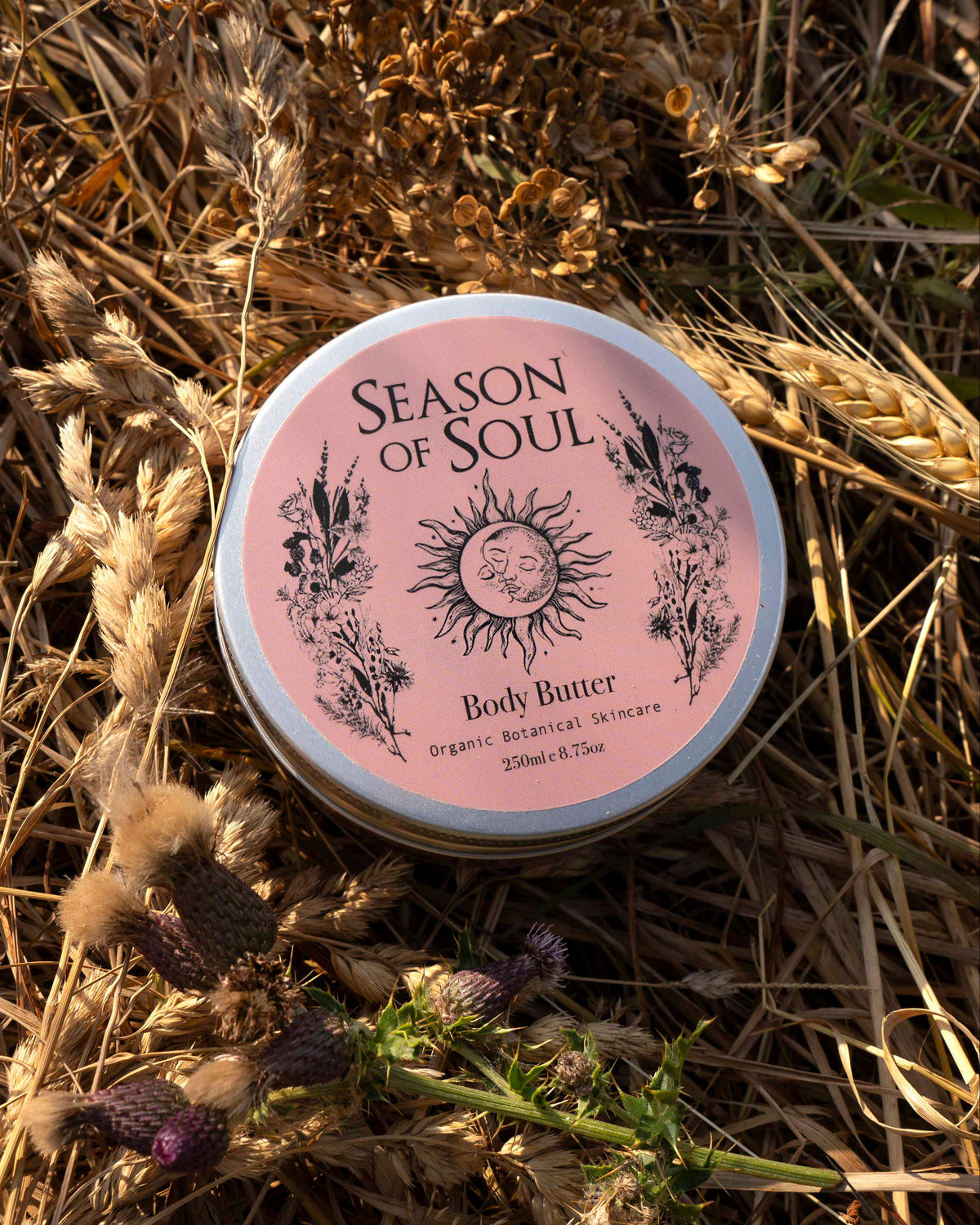 A round tin of Season of Soul Body Butter lies on a bed of dried wheat stalks and purple thistles. The lid is light pink with black text and illustrations of a sun and botanicals.
