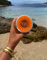 A hand holding a round tub of Season of Soul SPF 30 tinted moisturizer against a beach background with rocks and ocean water.